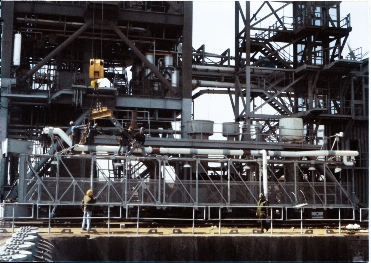 Image 100. At Space Shuttle Launch Complex 39-B, Kennedy Space Center, Florida, Union Ironworkers working for Ivey Steel Erectors, continue rigging the GOX Arm to lift it into place near the top of the Fixed Service Structure, which is visible directly behind them in this photograph taken from the east side of the Flame Trench. The west wall of the Flame Trench can be seen as the dark border along the bottom margin of the frame, and on its left side, the Spray Headers which lined the crest of the Flame Deflector can be seen. Our gang of four ironworkers is in the process of connecting the second spreader beam to the Arm Truss, and the one farthest left has his arm out, giving a signal to the crane operator to swing left with the Hammerhead Crane, which was used for this Lift. On the Pad Deck, Boeing TT&V personnel (who managed and operated the Hammerhead Crane now that it had become operational) can be seen, and the one on the left is relaying the hand signals to the crane operator up in the body of the crane at the control console (who cannot see any of what's going on directly) via walkie-talkie. The noticeable offset of the lifting gear, toward the left of the image, toward the Hinge end of the Arm, tells us that most of the weight of the Arm is in the Hinges, with the rest of it being quite light in comparison. On the far right end of the Arm, you can also see that the Vent Hood Assembly is not connected to the Arm, and it will be lifted and attached via a separate operation at a later time. Photo by James MacLaren.